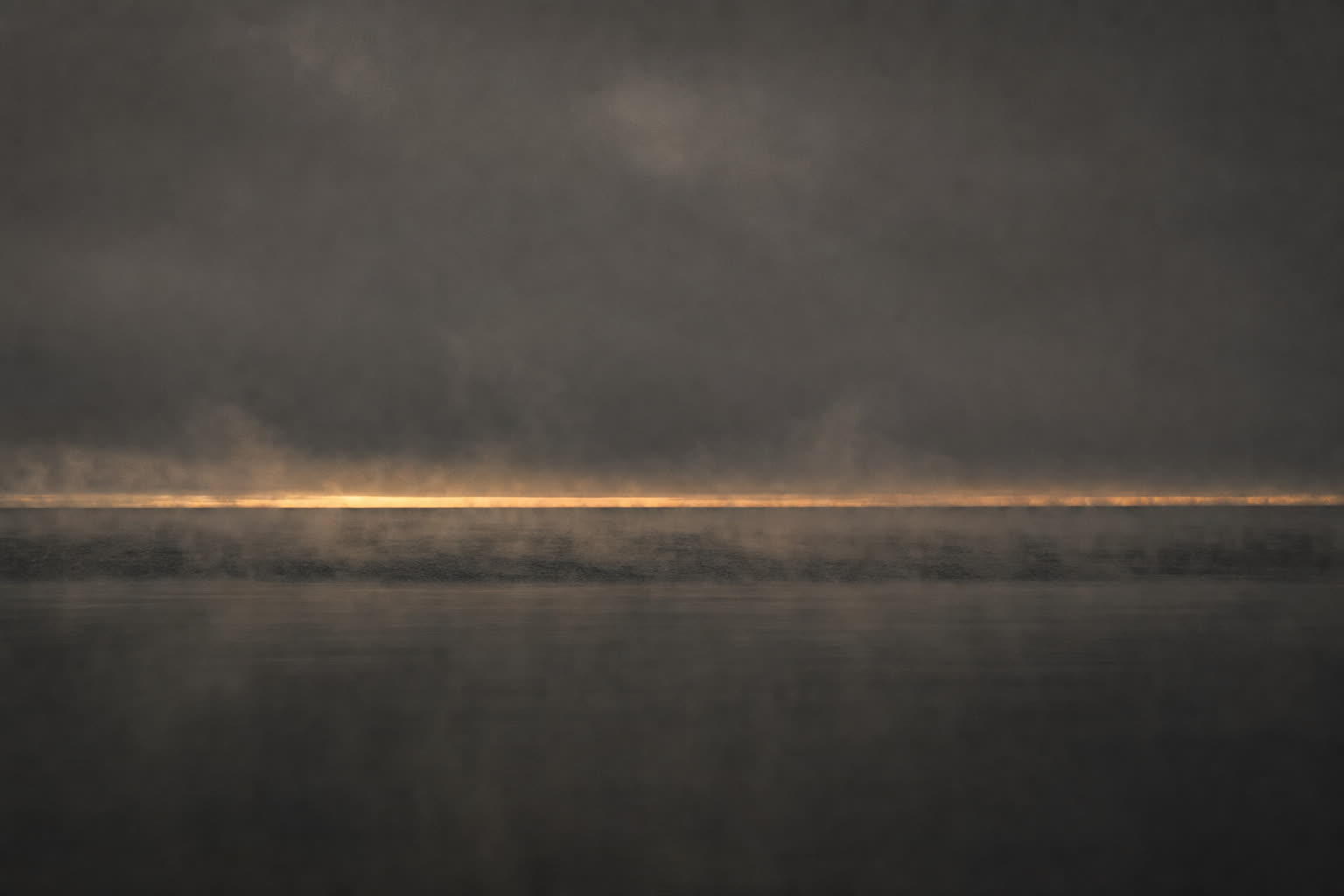 The surface of Lake Superior after a storm, calm black water with a thin band of amber horizon light.